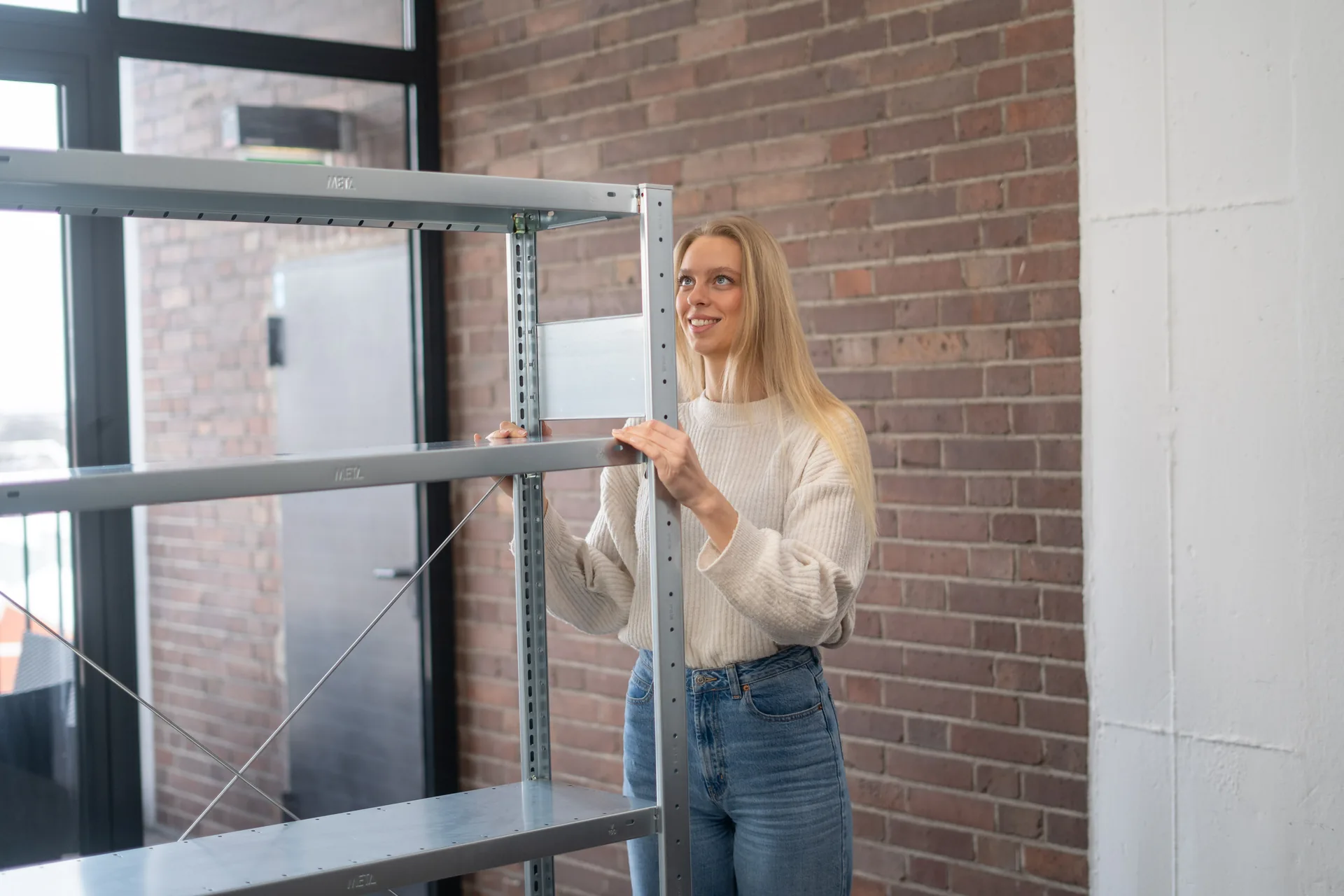 Frau baut ein verzinktes Metallregal vor einer Backsteinwand auf